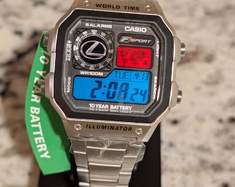 May include: A silver-tone Casio digital watch with a metal link band. The square face features a world time map, a Lexus logo, and a blue digital display showing 2:08:24 and the date 4-13. A green tag attached to the side reads 10 YEAR BATTERY.