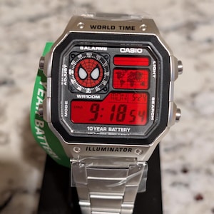 May include: A silver-tone digital watch with a metallic link band. The square face features a red background with a Spider-Man mask graphic, a world map display, and digital time reading 9:18. Text includes WORLD TIME, CASIO, and 10 YEAR BATTERY.