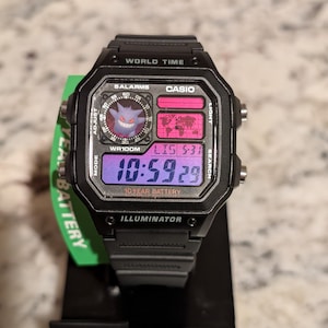 May include: A black Casio digital watch featuring a Gengar character graphic in the upper left dial, a world map display, and a digital time readout of 10:59:29. The watch face includes text labels for World Time, 5 Alarms, and 10 Year Battery.
