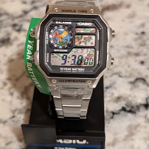 May include: A silver-tone Casio digital watch with a stainless steel band. The square face features a Bart Simpson graphic, a world map display, and digital text reading 9:38 PM, WED 3-24. The watch includes labels for World Time, 10 Year Battery, and Illuminator.