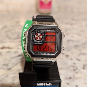 May include: A black and silver Casio digital watch with a red display showing the time. The watch has a black band and a green tag that reads "10 YEAR BATTERY". The watch is on a black stand.