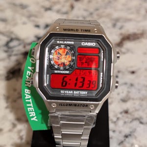 May include: A silver-tone Casio digital watch with a metallic link band. The rectangular face features a red digital display, a world map graphic, and a circular inset showing a Charizard character. Text includes WORLD TIME, 10 YEAR BATTERY, and ILLUMINATOR.