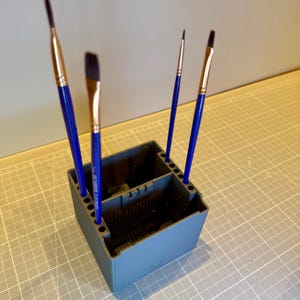 May include: A gray plastic brush holder with a square base and multiple circular slots along the top edges. Four paint brushes with blue handles and gold-toned ferrules are inserted into the slots, standing upright in the organizer.