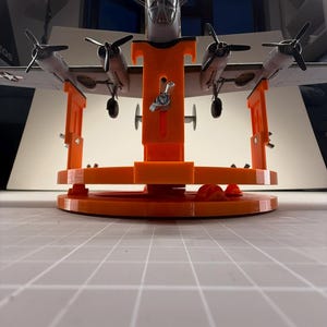 Könnte beinhalten: Ein leuchtend orangefarbener Modellflugzeugständer aus Kunststoff mit rundem Sockel und verstellbaren vertikalen Halterungen. Der Ständer verfügt über Metallflügelmuttern zur Höhenverstellung und trägt ein graues viermotoriges Modellflugzeug.