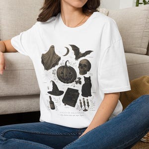 May include: A white short-sleeve t-shirt featuring a dark graphic print of Halloween-themed illustrations including a ghost, bat, crescent moon, pumpkin, skull, black cat, witch hat, spellbook, potion bottle, and candles with the text Spirits of Halloween.