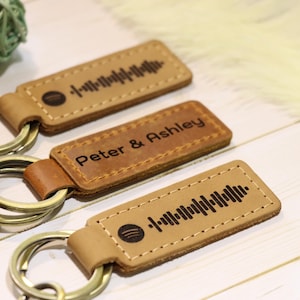 May include: Three leather keychains with metal rings. Two keychains feature a Spotify code design, and one reads "Peter & Ashley". The keychains are tan with brown accents and are on a light wood surface.