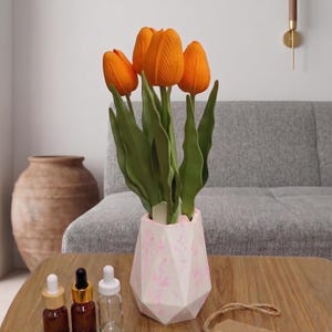 Könnte beinhalten: Ein Blumenarrangement mit orangefarbenen Tulpen in einer geometrischen Vase in Rosa und Weiß. Drei kleine braune Glasflaschen mit Tropfern und eine klare Flasche stehen auf einem Holztisch. Das Etikett lautet "AhfaAura".