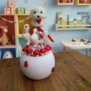 Puede incluir: Una pieza decorativa esférica blanca con acentos de hongos con tapa roja, adornos en forma de corazón y una figura de perro de peluche con un lazo rojo. La pieza está adornada con detalles de mariquitas y se asienta sobre una superficie de madera, con un tema lúdico.