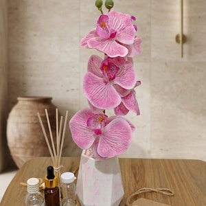May include: A pink orchid arrangement in a white and pink geometric vase. The image also shows small glass bottles, aroma diffuser sticks, and scented wax melts. A brown tag with the text "AhfaAura" hangs on the right.