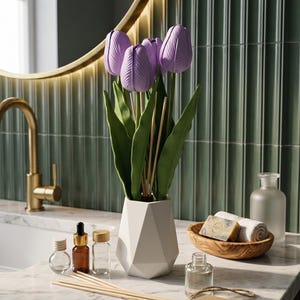 Puede incluir: Un jarrón geométrico blanco contiene tallos de tulipanes morados y hojas verdes. Varias botellas de vidrio pequeñas, palitos de madera y tabletas de cera perfumada están dispuestas sobre una encimera de mármol. También hay un cuenco de madera con toallas enrolladas y jabón. La etiqueta dice "AhfaAura".