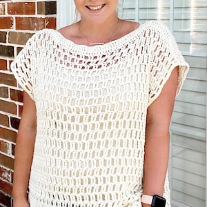 Pode incluir: Um top branco de crochê com um design solto e tecido aberto. O top tem um decote redondo e mangas curtas.