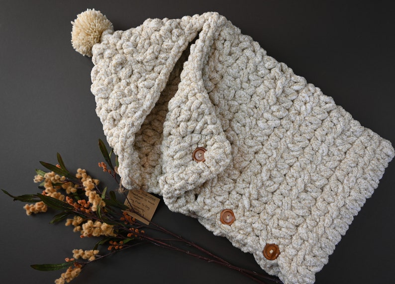 Pu&ograve; includere: Una coperta con cappuccio in crochet color crema con un pon pon sul cappuccio e bottoni in legno. La coperta &egrave; realizzata con un filato spesso e ha una texture morbida e soffice.