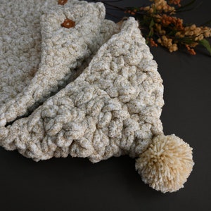 Pu&ograve; includere: Una sciarpa con cappuccio in crochet beige con un pompon soffice all'estremit&agrave;. La sciarpa &egrave; realizzata con un filato spesso e ha un aspetto testurizzato.