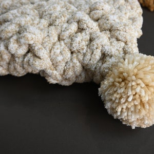 Pu&ograve; includere: Un berretto lavorato a uncinetto beige e bianco con un pompon soffice in cima.