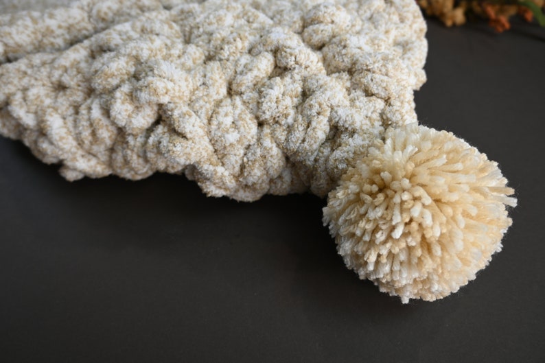 Pu&ograve; includere: Un berretto lavorato a uncinetto beige e bianco con un pompon soffice in cima.