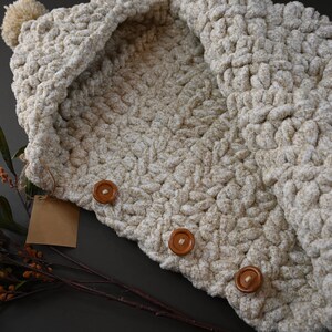Pu&ograve; includere: Una sciarpa con cappuccio in crochet color crema con un pon pon in cima e tre bottoni in legno marrone.