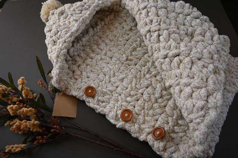 Pu&ograve; includere: Una sciarpa con cappuccio in crochet color crema con un pon pon in cima e tre bottoni in legno marrone.