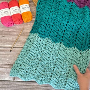 Könnte beinhalten: Eine teal-, aqua- und lilafarbene Häkeldecke mit einem gewellten Rand. Die Decke ist mit einem zarten Lochmuster gearbeitet und wird auf einer Holzoberfläche mit Garnknäueln gezeigt.