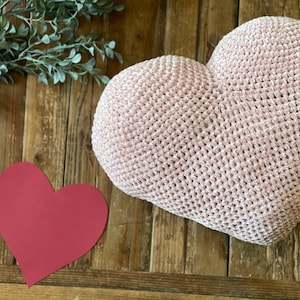 May include: A pink crocheted heart-shaped cushion on a wooden surface. A red paper heart is in the foreground.