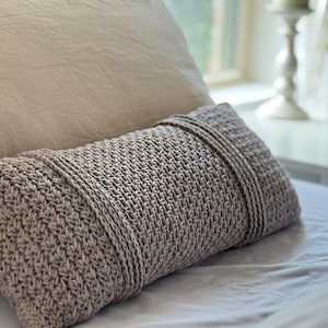 Può includere: Un cuscino rettangolare all'uncinetto color talpa con un motivo a punto strutturato. Il cuscino ha un dettaglio a coste sui bordi superiore e inferiore. Poggia su un letto con un cuscino e un lenzuolo di colore chiaro.