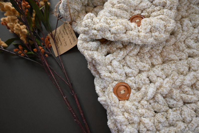 Pu&ograve; includere: Una coperta all'uncinetto color crema con due bottoni in legno. La coperta &egrave; realizzata con un filato spesso e ha una texture morbida e soffice.