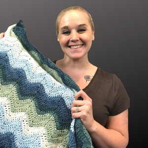 May include: A person holding a crocheted blanket with a chevron pattern in shades of blue, green, and white.