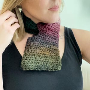 May include: A crocheted neck warmer in shades of green, purple, and brown. The neck warmer is made with a chunky yarn and has a unique asymmetrical shape.
