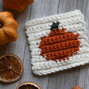 May include: A white crocheted square with an orange pumpkin design in the center. The pumpkin has a green stem and is surrounded by white crocheted stitches.