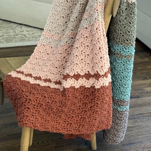 May include: A crocheted throw blanket with a geometric pattern in shades of brown, pink, and teal. The blanket is draped over a wooden chair.