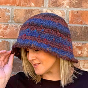 May include: A handmade crochet bucket hat in shades of blue, brown, and red. The hat features a striped pattern and a slightly downward-sloping brim. The hat is made of a textured yarn.