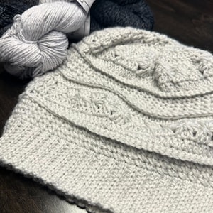 May include: A close-up of a crocheted hat in a light gray color. The hat has a textured design with multiple rows of stitches.