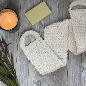 May include: A white crocheted exfoliating body scrub with a loop handle. The scrub is on a wooden surface with a candle and a bar of soap.