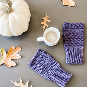May include: A pair of purple crocheted fingerless gloves with a white pumpkin, a cup of coffee, and fall leaves on a gray surface.