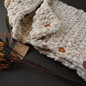 Pu&ograve; includere: Una sciarpa con cappuccio color crema realizzata a uncinetto con bottoni in legno marrone. La sciarpa &egrave; realizzata con un filato morbido e soffice e presenta un motivo intrecciato testurizzato. La sciarpa &egrave; appoggiata su una superficie grigio scuro con un rametto di fiori secchi.