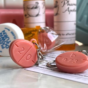 May include: Two coral-colored pill-shaped keychains with silver keyrings. One keychain has the number "20" and the other has "b 973" imprinted on it. The keychains are in front of pill bottles and a prescription.
