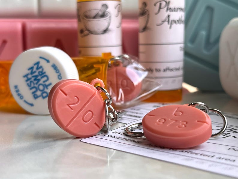 May include: Two coral-colored pill-shaped keychains with silver keyrings. One keychain has the number "20" and the other has "b 973" imprinted on it. The keychains are in front of pill bottles and a prescription.