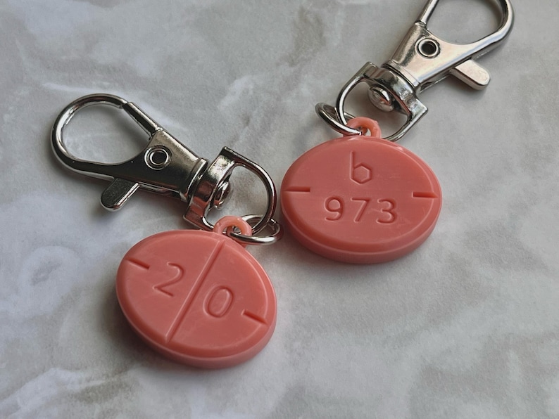 May include: Two pink pill-shaped keychains with silver-colored clasps. One keychain has the number "20" and the other has "973" and the letter "b" imprinted on it. The keychains are on a light-colored surface.