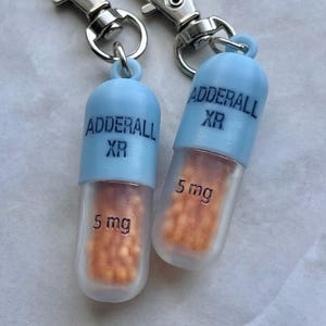 Può includere: Due portachiavi a forma di pillola, azzurri e trasparenti, con chiusure argentate. I portachiavi sono etichettati "ADDERALL XR" e "5 mg". La parte trasparente contiene perline arancioni.