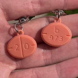 May include: A pair of peach-colored earrings shaped like pills. One earring has the number "20" and the other has "b 973" embossed on the surface. The earrings are attached to silver-colored metal hooks.