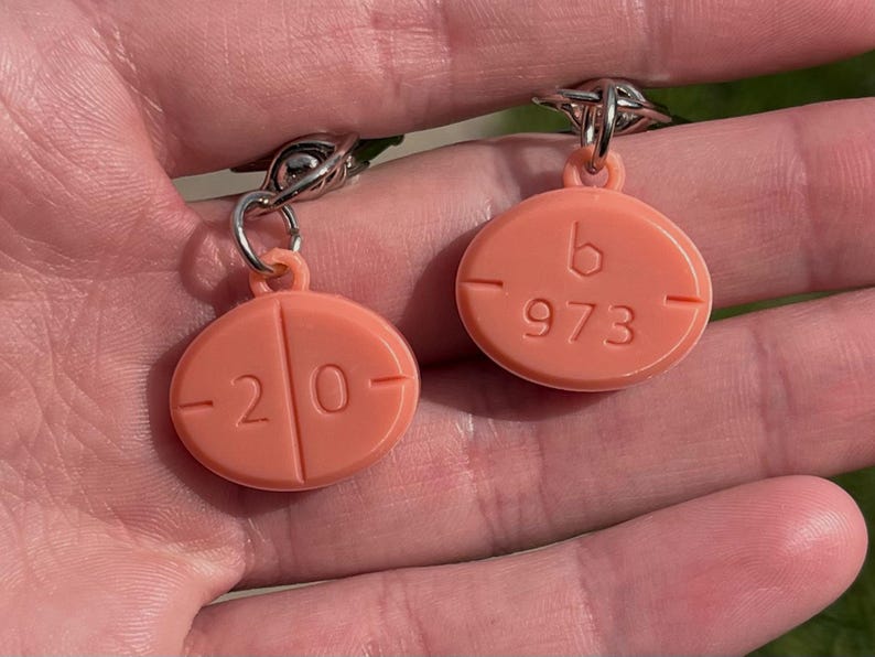 May include: A pair of peach-colored earrings shaped like pills. One earring has the number "20" and the other has "b 973" embossed on the surface. The earrings are attached to silver-colored metal hooks.