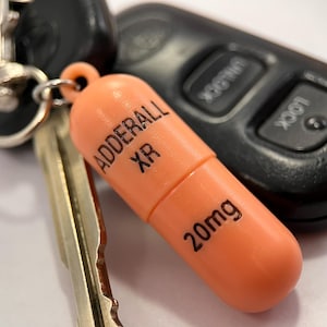 Op de afbeelding: Een perzikkleurige, pilvormige sleutelhanger met de zwarte opdruk ADDERALL XR en 20mg. De hanger is bevestigd aan een zilverkleurige metalen ring die verbonden is met een zwarte autosleutel en een metalen sleutel, wat een functioneel accessoire vormt.