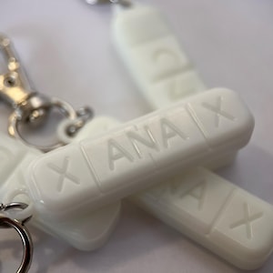 以下が含まれることがあります： 角が丸い白い長方形のキーホルダーで、両面にエンボス加工されたANAXの文字とXが刻まれています。キーホルダーには銀色の金属製ロブスタークラスプと丸いスプリットリングが付いており、バッグや鍵に取り付けられるように設計されています。