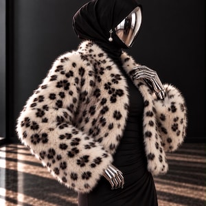May include: A cropped jacket featuring a cream-coloured base with a dark brown leopard print pattern. The jacket has long sleeves and a high collar. It is worn over a plain black dress. The mannequin has a black head covering and a silver face.