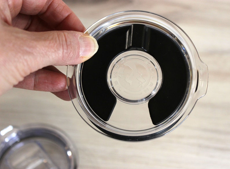 May include: A clear plastic lid with a black rubber seal and a small silver logo. The lid has a lip around the edge and a small handle on the side.