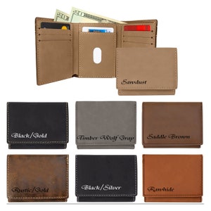 May include: A collection of trifold wallets in various colors including black, gray, brown, and rustic tan. Each wallet features visible stitching and engraved text labels such as Black/Gold, Timber Wolf Gray, Saddle Brown, Rustic/Gold, Black/Silver, and Rawhide.