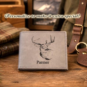 May include: A gray bi-fold wallet featuring a black laser-engraved illustration of a deer head and the name Parmer centered below the image. The wallet has visible stitching along the edges and a smooth, matte finish on the front surface.