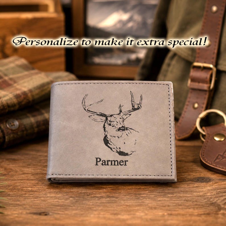 May include: A gray bi-fold wallet featuring a black laser-engraved illustration of a deer head and the name Parmer centered below the image. The wallet has visible stitching along the edges and a smooth, matte finish on the front surface.