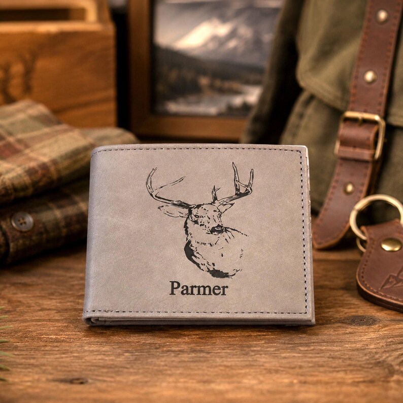 May include: A gray bi-fold wallet featuring a black engraved illustration of a deer head centered on the front cover. Below the deer, the name Parmer is printed in a black serif font. The wallet has visible stitching along the edges.