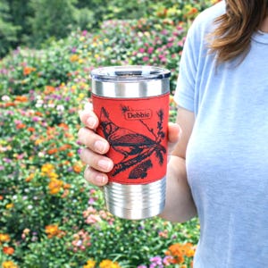 May include: A stainless steel travel mug with a red leather wrap featuring a black cardinal bird design and the name "Debbie". The mug has a silver lid and base, perfect for hot or cold beverages.