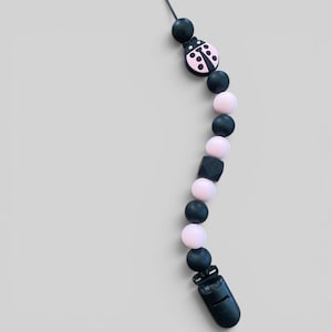 May include: A black and pink baby pacifier clip with a ladybug design. The clip features a series of black and pink beads, a black clip, and a ladybug-shaped charm. The pacifier clip is designed to attach to a baby's clothing.